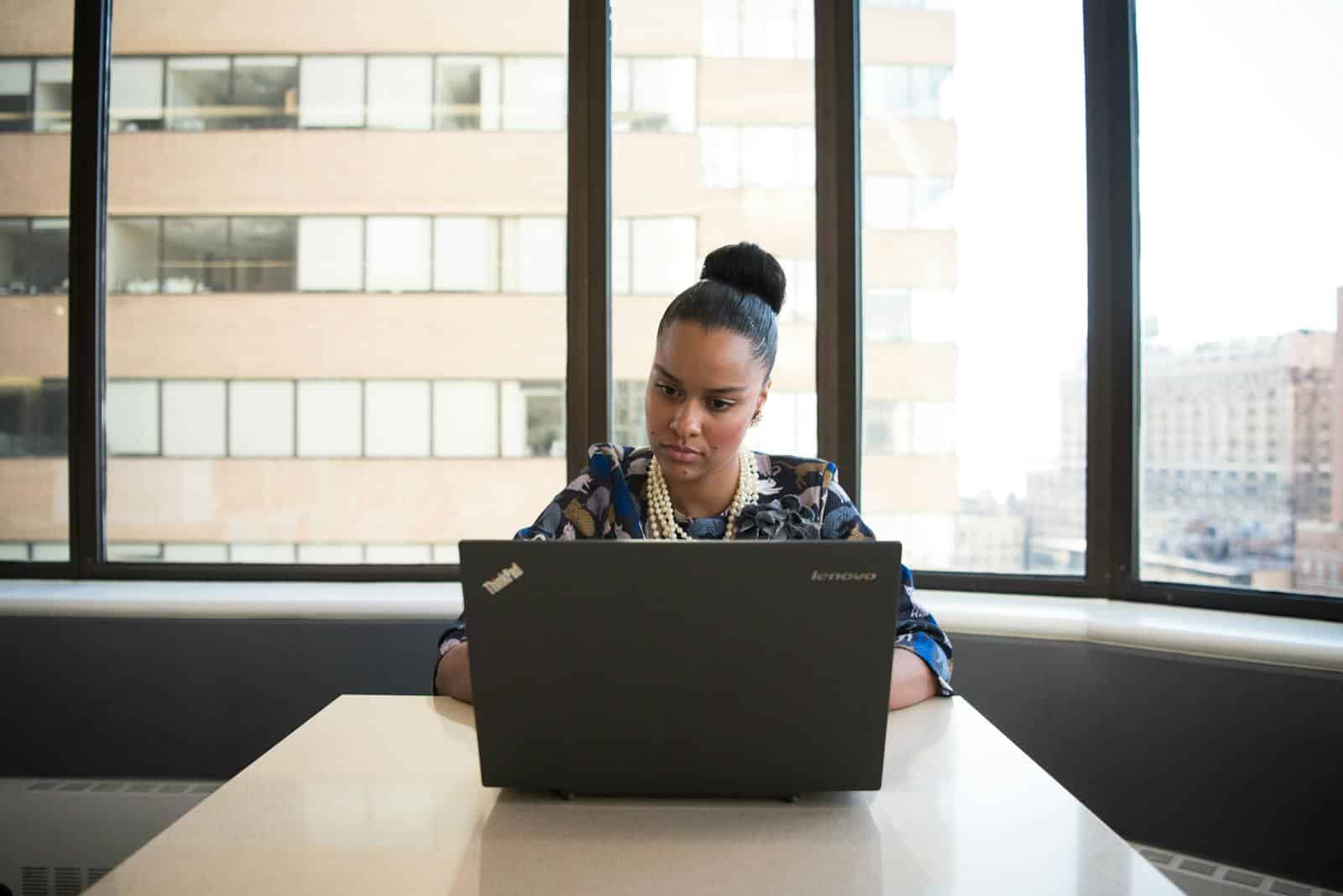 Professional woman using a laptop at a desk in a modern office setting with city views.